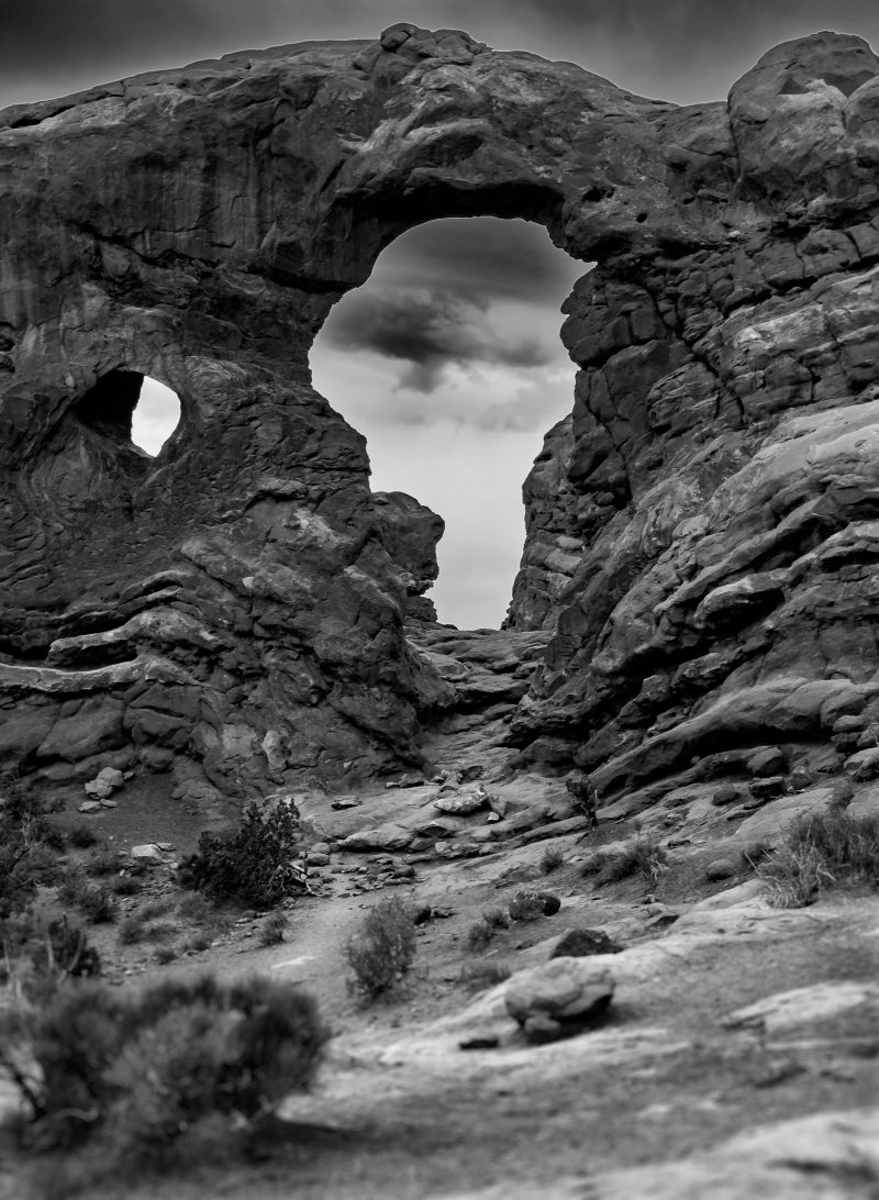 A black and white photograph of a sandstone arch with a smaller window in the rock, desert scrub beyond