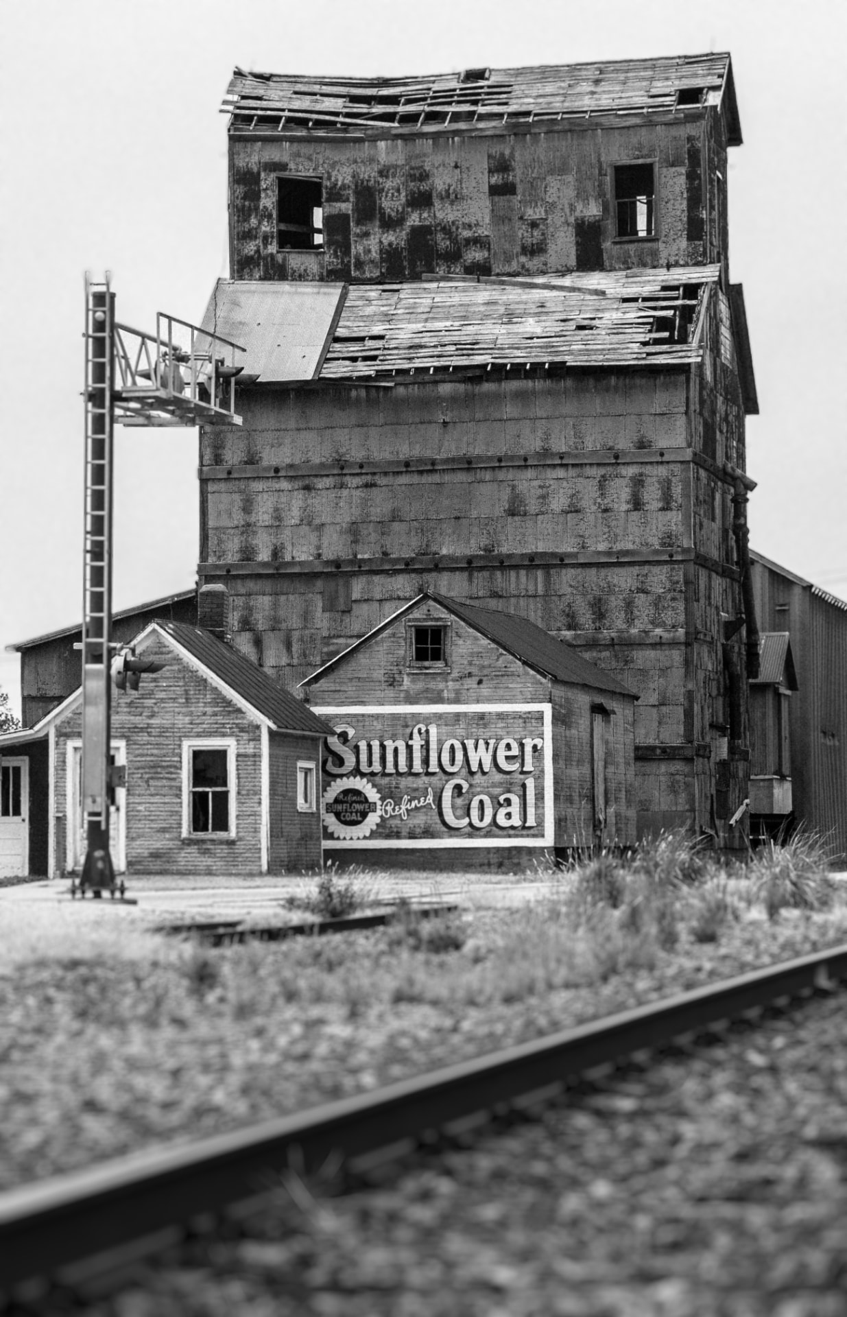 A black and white portrait view of the derelict Sunflower Coal grain elevator and signal gantry by the tracks