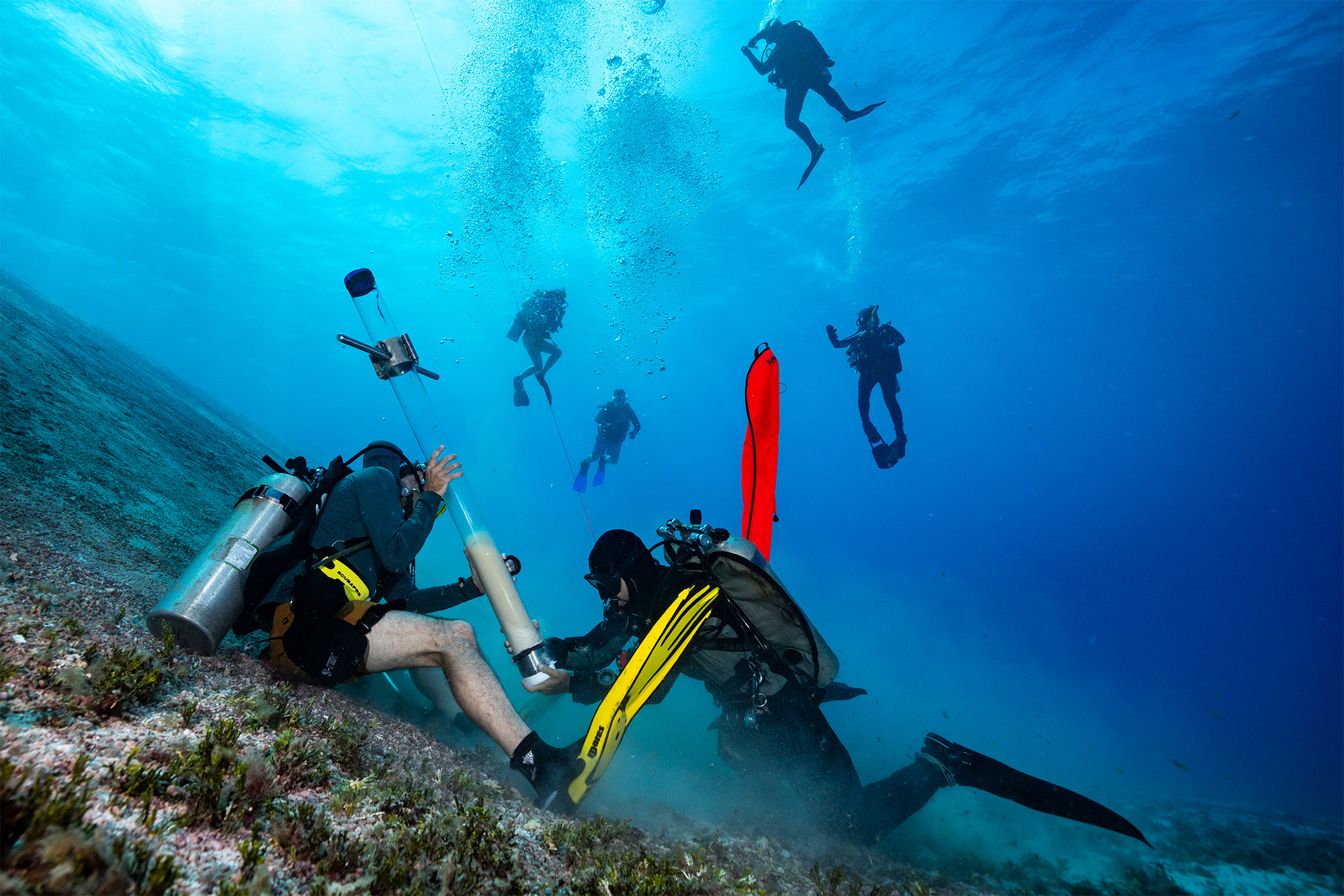 Divers driving a core tube into the reef slope while the team hangs above in blue water