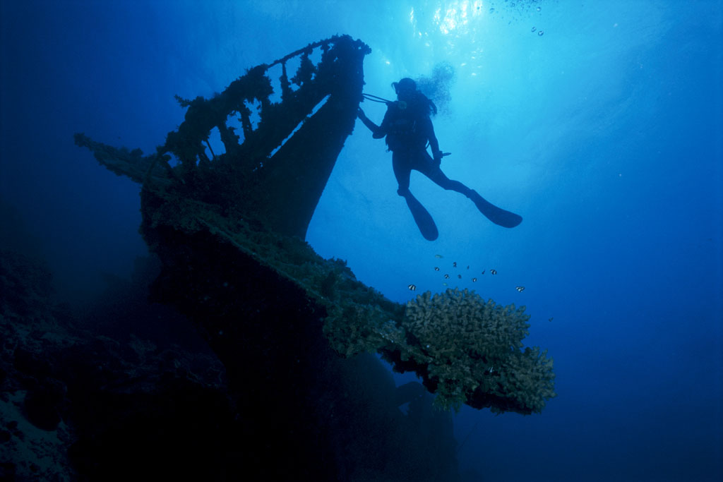 A diver silhouetted against blue water above a coral covered wreck