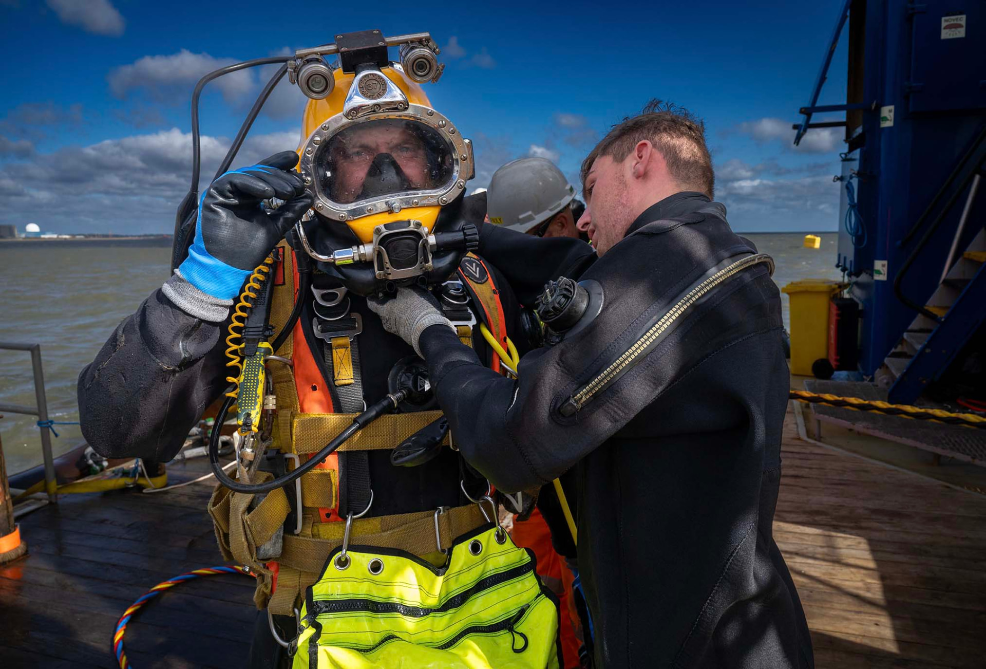 A tender helping a commercial diver secure his helmet on deck